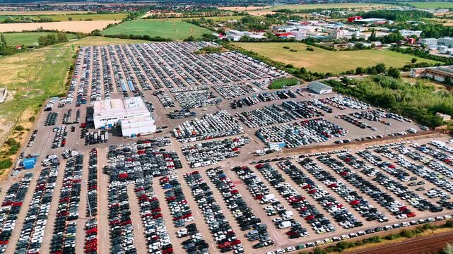 A car storage facility spreads across a paved site. Thousands of vehicle roofs form a grid around a central building with fields and houses beyond.