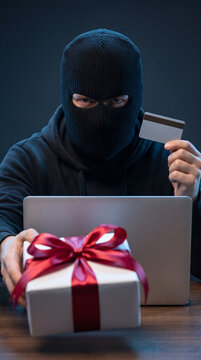 Masked cybercriminal holding credit card behind laptop and wrapped christmas gift representing online shopping fraud.