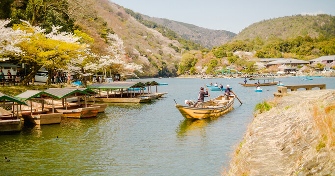 Kyoto, Kyoto Prefecture, Japan - 10 April 2025: View of Arashiyama with traditional wooden boats on the Hozugawa River, surrounded by blooming cherry blossoms and lush hills under a clear sky.