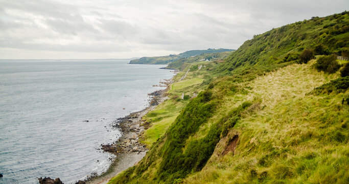 Cushendun, County Antrim, United Kingdom - 14 September 2024: View of the rugged coastline with green grassy cliffs sloping down to the sea under a cloudy sky.