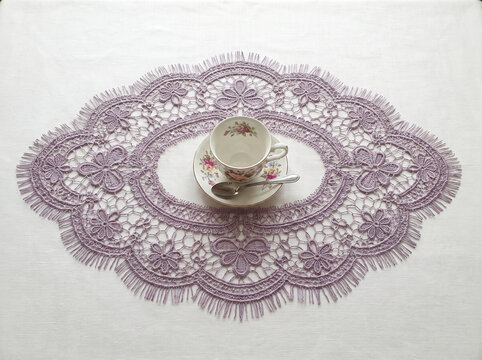 Lavender lace doily mat and elegant floral teacup with saucer and spoon on a white linen table surface, ideal for home decor, craft, and vintage design concepts.