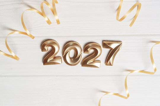 Shiny golden 2027 numbers centered between curled gold ribbons on white painted wooden planks
