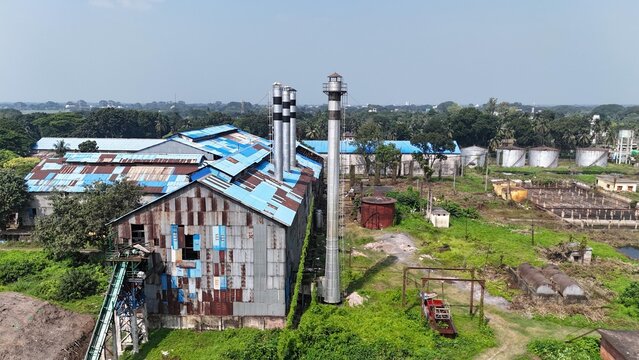 Aerial view of a rural industrial factory with corrugated metal walls, blue tin roofs, and tall steel chimneys surrounded by green vegetation