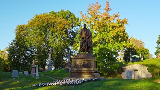 A statue stands above a cemetery pedestal in Brooklyn. A robed figure faces the viewer beside a low tomb with rows of small lights across the grass.
