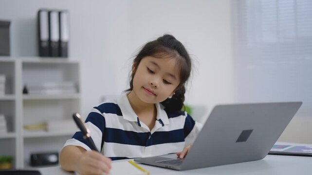Young girl sitting at desk using laptop and writing with pen in notebook focused on learning indoors