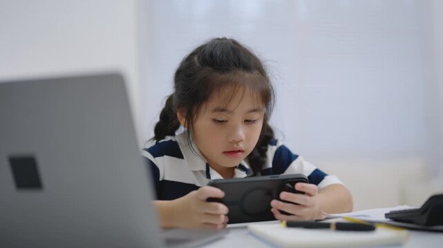 Young girl focused on handheld device screen while sitting at desk near laptop with notebook and pen indoors