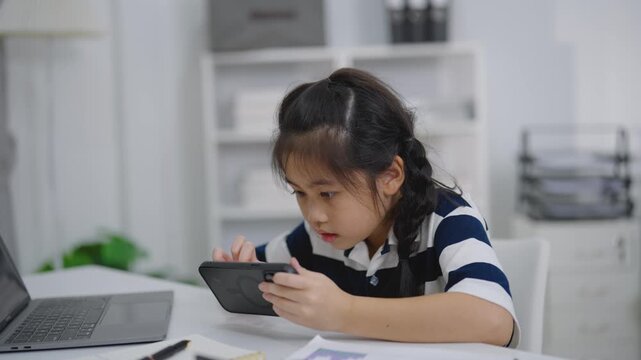Young girl playing game on mobile phone with intense focus at home desk beside laptop in casual environment
