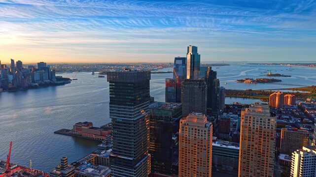 Jersey City towers overlook the Hudson River and harbor. Dark glass facades rise above sunlit buildings with blue water and distant islands beyond.