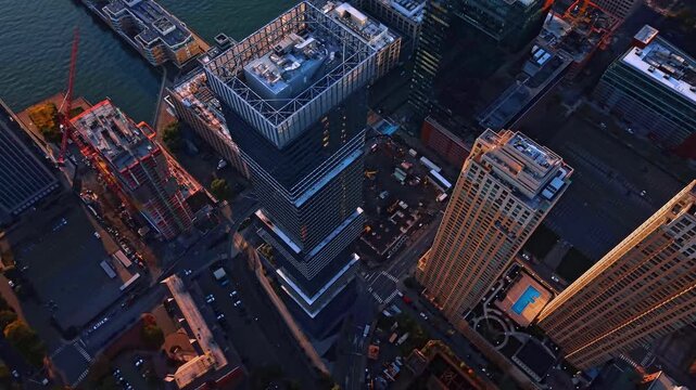 A stepped glass tower rises beside the Jersey City waterfront. Rooftop equipment and construction cranes appear below the steep viewpoint with a blue pool at the right.