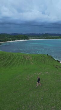 Woman Walking On Grassy Hill Above Rolling Ocean, Solitary Hiker Exploring Coastal Cliffs At Dawn, Narrow Dirt Path Winding Toward Shoreline, Bukit Merese, Lombok, Tanjung Ann, vertical video