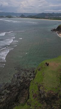 Aerial Seaview With group of people On Cliff Overlooking Turquoise Ocean And Sandy Bay Two Travelers Sit On Grassy Promontory Above Rugged Rocks, Gentle Waves Rolling Into Shoreline, Lombok