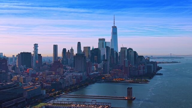 One World Trade Center rises above Lower Manhattan. Dense waterfront buildings border blue harbor water beneath thin bands of pink and white clouds.
