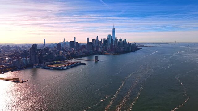 Lower Manhattan borders the Hudson River beneath streaked clouds. A spired tower rises near the center while boat wakes curve across water beside projecting piers.