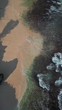 Shimmering emerald seascape with vibrant plant life surrounding rugged shoreline. Bright green waters swirl vigorously around jagged rocks amid abundant plant coverage, vertical video of Bali coast