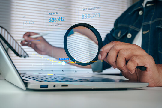 Businessman holding magnifying glass to analyze digital financial dashboard chart with ROI, net profit, and investment metrics on laptop screen, concept of financial audit and analysis.
