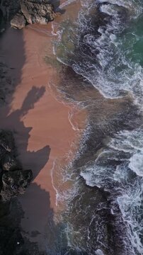 Waves crash shore. Crashing ocean waves swell over coral pink sand and rugged stones. Foamy waves breaking over vibrant pink sands and sharp rocky formations along coastline, Uluwatu, Bali, Indonesia