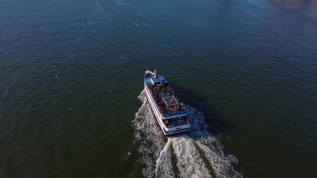 A passenger boat travels away across the Hudson River. Rows of people fill the open upper deck above a white stern and a foamy trail viewed from behind.