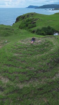 Woman Reclines On Cliff Beside Ocean, Solo Traveler Enjoying Panoramic Aerial View Of Turquoise Bay And Sandy Cove, Green Hills And Winding Shore In Background, Loyal Dog Curled Nearby, Lombok