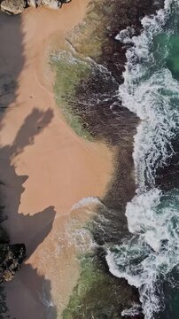 Crashing surf meets sandy shoreline. Foamy waves clash with rugged rocks near sandy coast. Rough stones endure relentless surf as pink sands shimmer beneath gloomy sky,Bali,Indonesia,vertical video