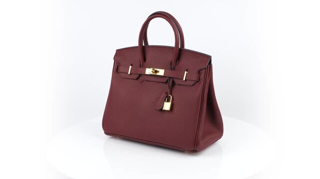 Seamless 360 degree rotation of a burgundy handbag in 4K studio footage, showing its pebbled texture, top handle, metal clasp and lock on a clean white background.