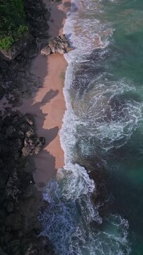 Waves crash shore. Crashing ocean waves swell over coral pink sand and rugged stones. Foamy waves breaking over vibrant pink sands and sharp rocky formations along coastline, Uluwatu, Bali, Indonesia