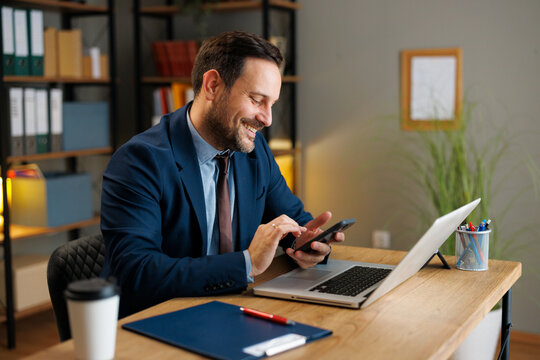 Smiling businessman using smartphone beside laptop at office desk