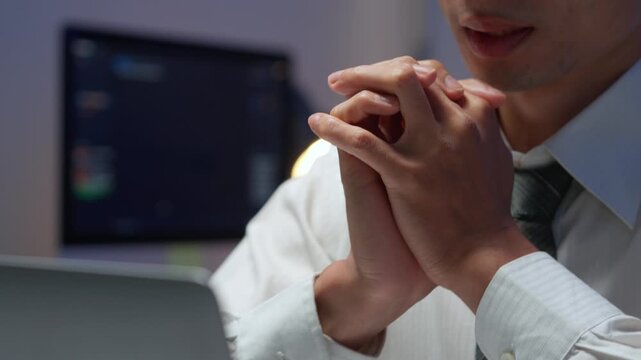 Anxious asian programmer praying with clasped hands while waiting for software deployment results late at office desk