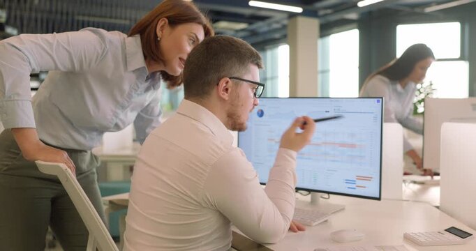 Manager and analyst review budget data on computer. A businesswoman leans over a young colleague who points at a budget variance report on the screen. Mentoring and financial control at work.