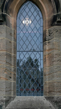 An arrow-shaped (lance-shaped) stained-glass window with a diamond- or rhombus-shaped pattern (grid), set into the stone wall of the church