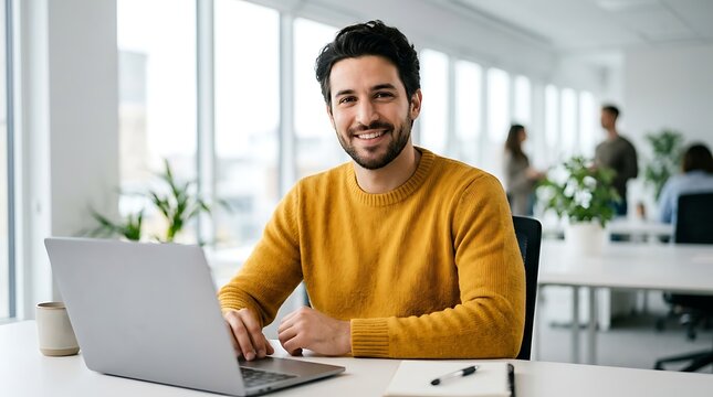 Confident businessman working on laptop computer at office desk, smiling professional man in casual yellow sweater, modern corporate workplace environment