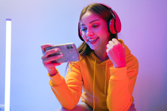 Young woman in headphones smiles while using a smartphone in colorful studio lighting
