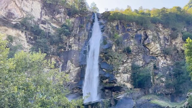 Majestic waterfall cascading down steep rocky cliff surrounded by lush forest in Foroglio, Switzerland, scenic alpine nature landscape on sunny day