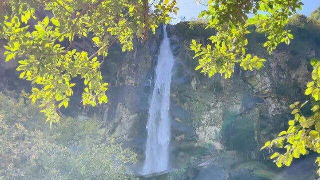 Tall waterfall cascading down a rocky cliff framed by sunlit green leaves in Foroglio, Switzerland, scenic alpine nature landscape