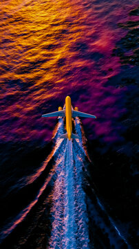 A yellow airplane is flying over a body of water with a colorful sunset in the background