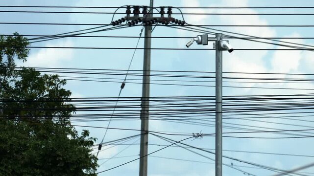 Messy grid of black wires and security cameras on utility poles against bright sky