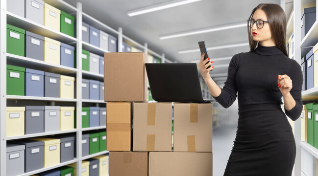Archive management. Female employee uses a smartphone and laptop to manage inventory, organize stored documents and track cardboard boxes in a records storage facility.
