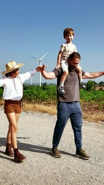 Parents walk along a rural gravel path with their young child riding on his father's shoulders beside wind turbines