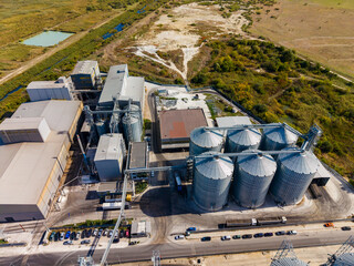 Aerial view of an industrial grain processing facility with storage silos beside a road and open land © DSMT
