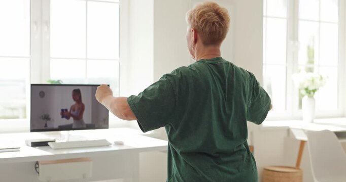 Senior woman follows online workout fitness training. Back view footage shows her extending arms while watching instructor on computer in bright home. Digital wellness supports active aging.