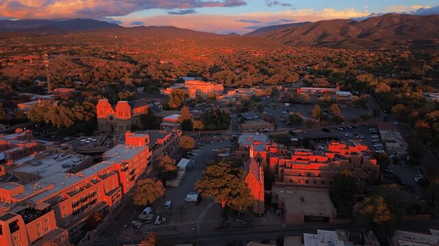 San Loretto chapel Santa Fe, capital of New Mexico, USA glows red in last sun beside Cathedral Basilica and adobe hotels. Aerial orbit over autumn trees, parking lots and dark mountains at sunset.