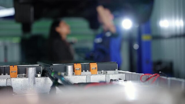 Sharp foreground of an open EV battery pack with orange connectors and red wires while a blurred woman and mechanic point up at the underside of a car on the lift behind