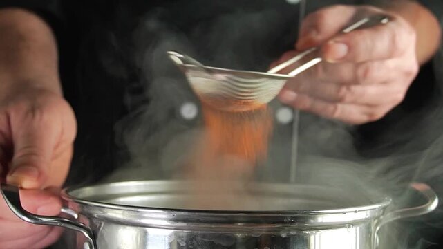 Hands sift orange spice through a fine strainer into a steaming stainless steel pot during cooking, showing close culinary preparation in a dark kitchen setting