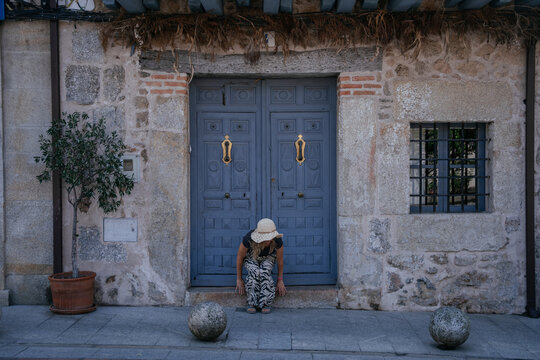 Middle Aged European Female Resting on Doorstep of Traditional Stone House with Blue Wooden Doors in Spanish Village