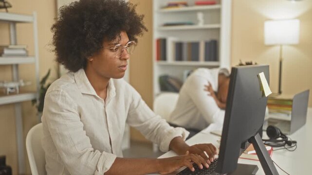 Man at desktop computer with glasses and afro, hand covering mouth while typing and yawning in office building with coworker slumped at desk; tired focus.