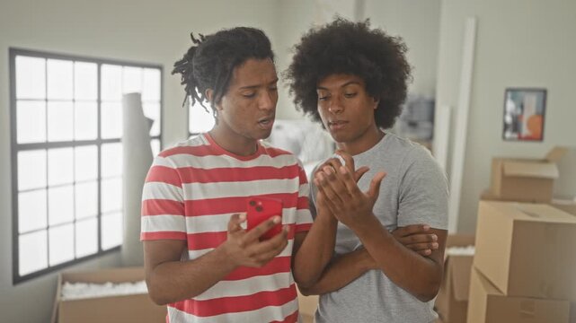 Two men pointing finger to smartphone while checking a checklist amid stacked moving boxes in a sunlit building with large window; focus teamwork.
