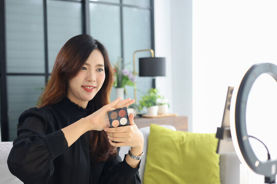 Beautiful Asian female beauty blogger live streaming makeup tutorial at home holding eyeshadow palette and brush in front of ring light.
