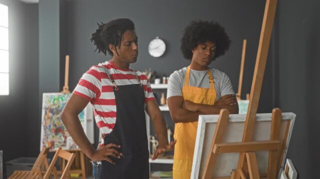 Two men painters wearing aprons point to canvas on easel with hands discussing composition in art studio; creative focus.