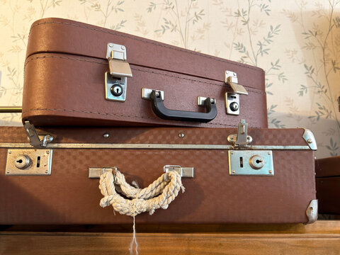 two weathered brown vintage suitcases stacked against floral wallpaper, antique travel luggage with metal fasteners and rustic rope handle