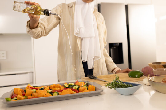 Adult African American woman pouring olive oil from glass bottle onto rimmed baking sheet with veg