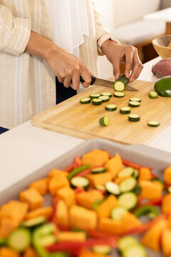 African American woman slicing zucchini using chef knife on board on white counter wearing watch